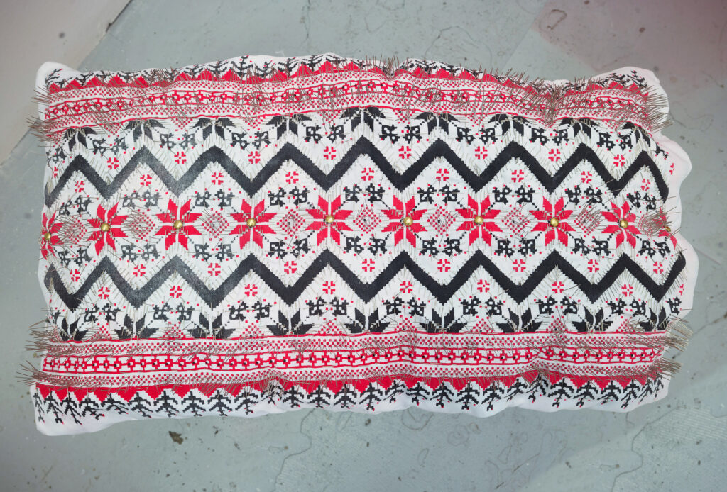 The artwork "A Pillow" embodies life's duality, drawing inspiration from contrasting experiences of celebration and exposure to ongoing violence through social media and news. It captures the juxtaposition of witnessing New Year's fireworks alongside the stark realities of war in Gaza. The piece features a traditional Palestinian embroidery pattern screen-printed on a white burial shroud, meticulously crafted with hundreds of sewing needles pointing outward, later stitched into a pillow. The transformation of the pillow with sharp metallic pins represents discomfort and pain, subverting its role as a symbol of comfort and rest. It symbolizes mental and physical exhaustion, particularly during restless and sleepless nights. Embedded within the pillow is a speaker playing an audio composition I've created, blending recorded sounds of New Year's fireworks with missiles dropping in Gaza on the same night.